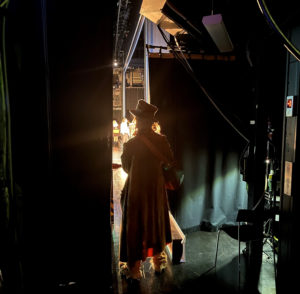 Daniel Lemma står, med ryggen mot kameran, precis vid scenöppningen iklädd hatt och med en gitarrem över axeln. Ljuset från scen lyser mot kameralinsen och skådespelarna skymtar i bakgrunden.